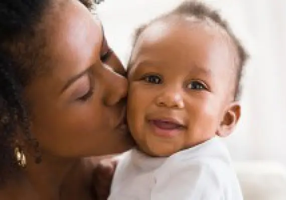 A woman lovingly kisses a smiling baby on the cheek.