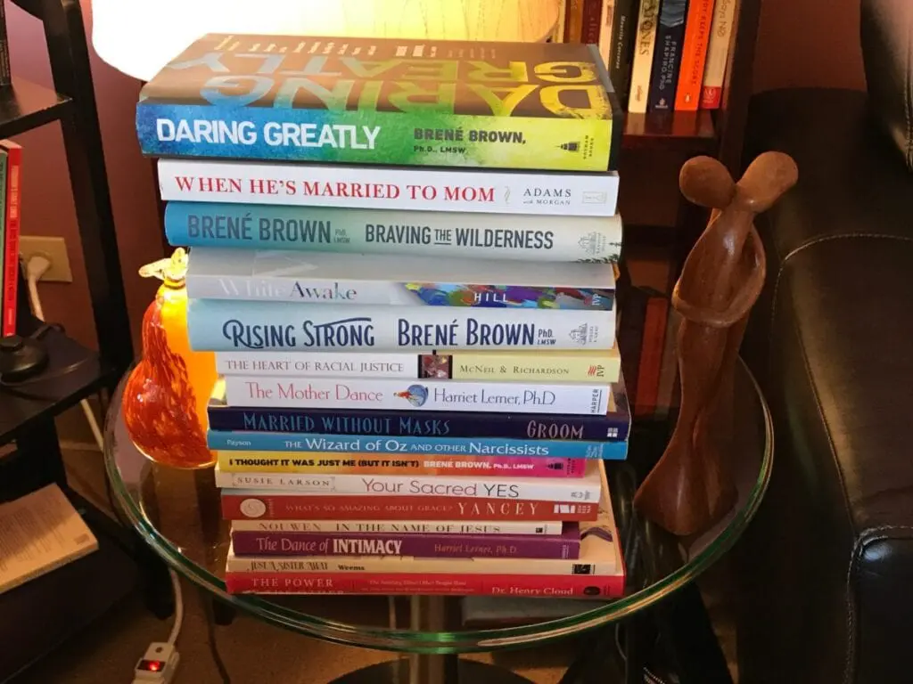 A stack of various self-help and relationship books is on a glass table, next to an abstract wooden sculpture.