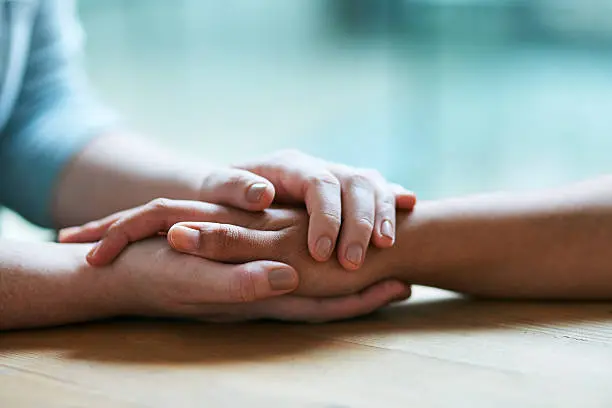 Two pairs of hands are gently holding each other on a table, symbolizing support and understanding.