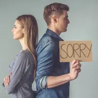 A man holding a "Sorry" sign stands back-to-back with a woman, both looking away from each other.