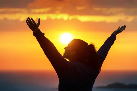 A person with outstretched arms stands before a vibrant sunset or sunrise over the ocean.