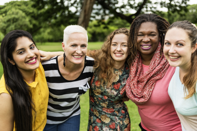 A group of five women smiling and posing together outdoors, possibly in a park setting.