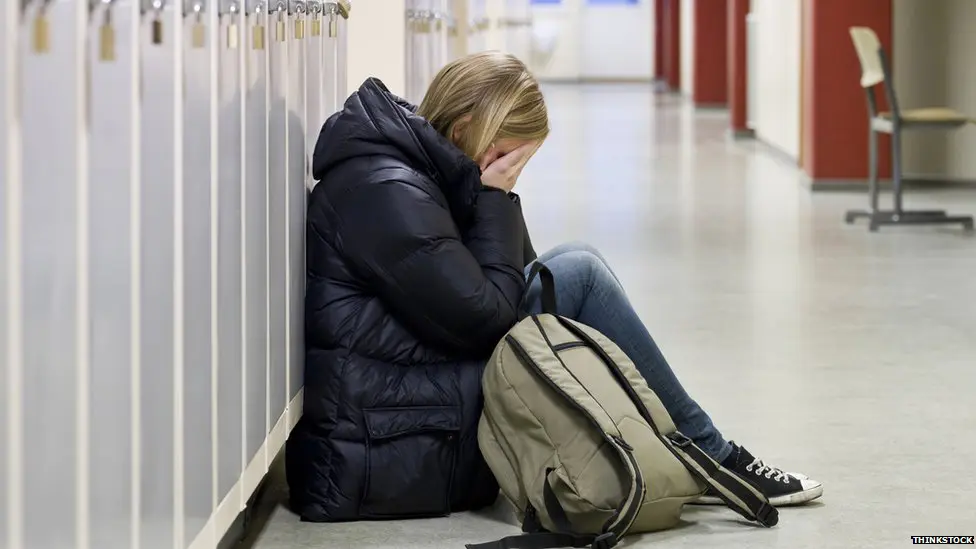 A person sitting on the floor in a hallway, holding their head in their hands, next to a backpack.
