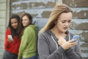 A girl looks upset while holding her phone, with two others in the background looking at another phone.