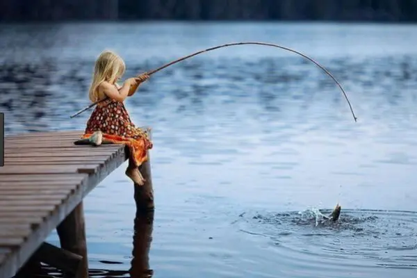 A young girl sits on a dock, fishing with a simple rod, as a fish splashes in the water.