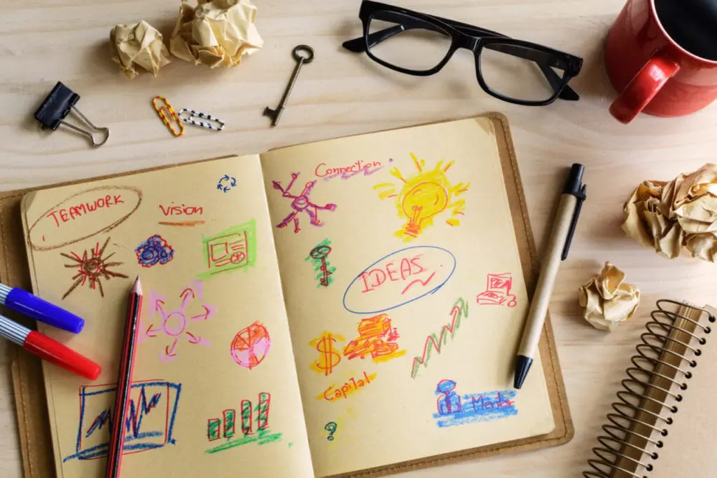 A notebook with colorful sketches and words like "TEAMWORK," "Vision," "IDEAS." Nearby are pens, glasses, crumpled papers, and a mug.