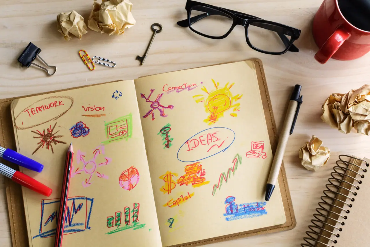 A notebook with colorful sketches and words like "TEAMWORK," "Vision," "IDEAS." Nearby are pens, glasses, crumpled papers, and a mug.