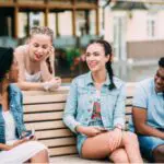 A group of friends is sitting and chatting on a bench outdoors, enjoying a relaxed moment.