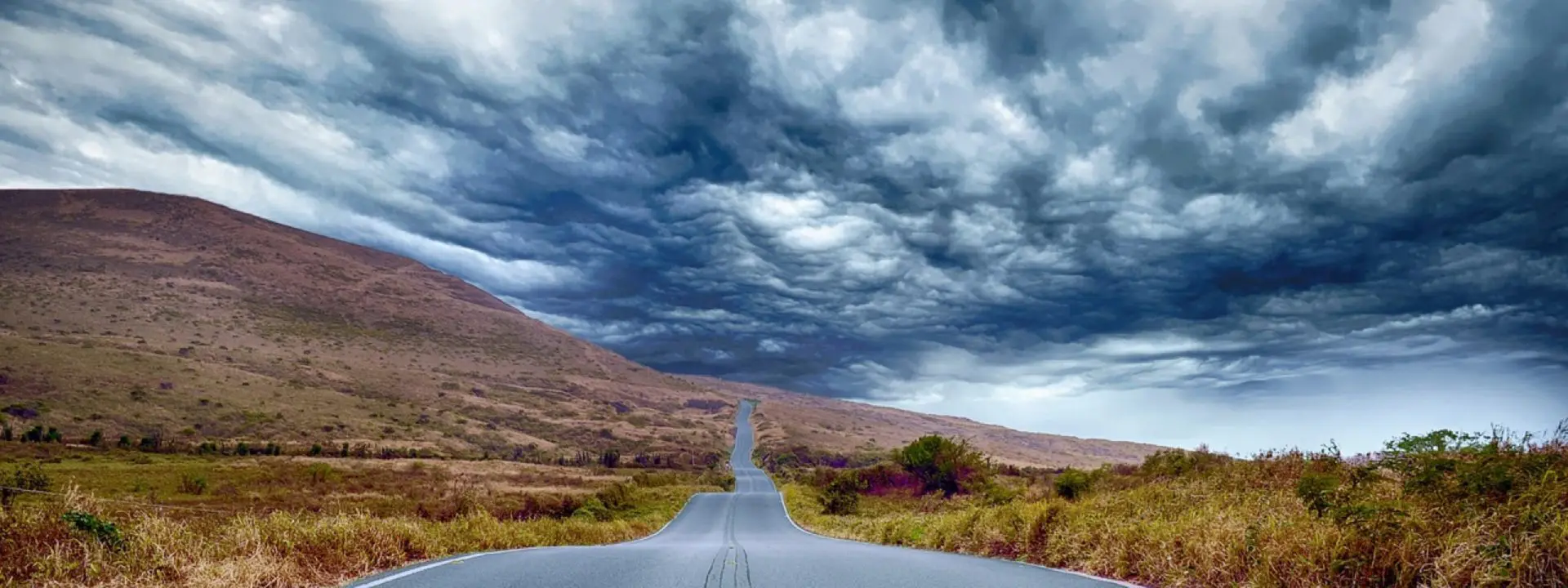 A winding road stretches into the distance with dramatic storm clouds overhead, flanked by hills and grassland.