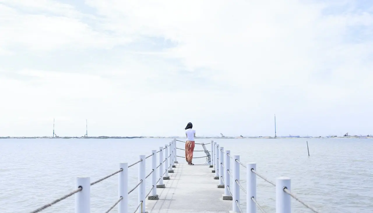 A person stands alone on a pier overlooking a calm sea under a bright sky, creating a serene and contemplative scene.