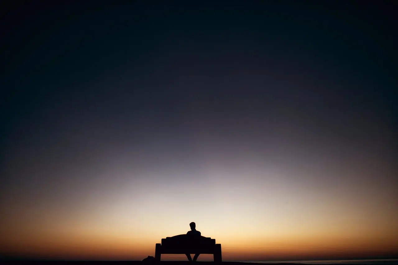 A person sits on a bench gazing at a peaceful sunrise or sunset, evoking tranquility and contemplation.