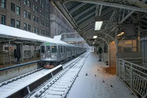 A snowy train platform with a train arriving, surrounded by buildings and a few pigeons.