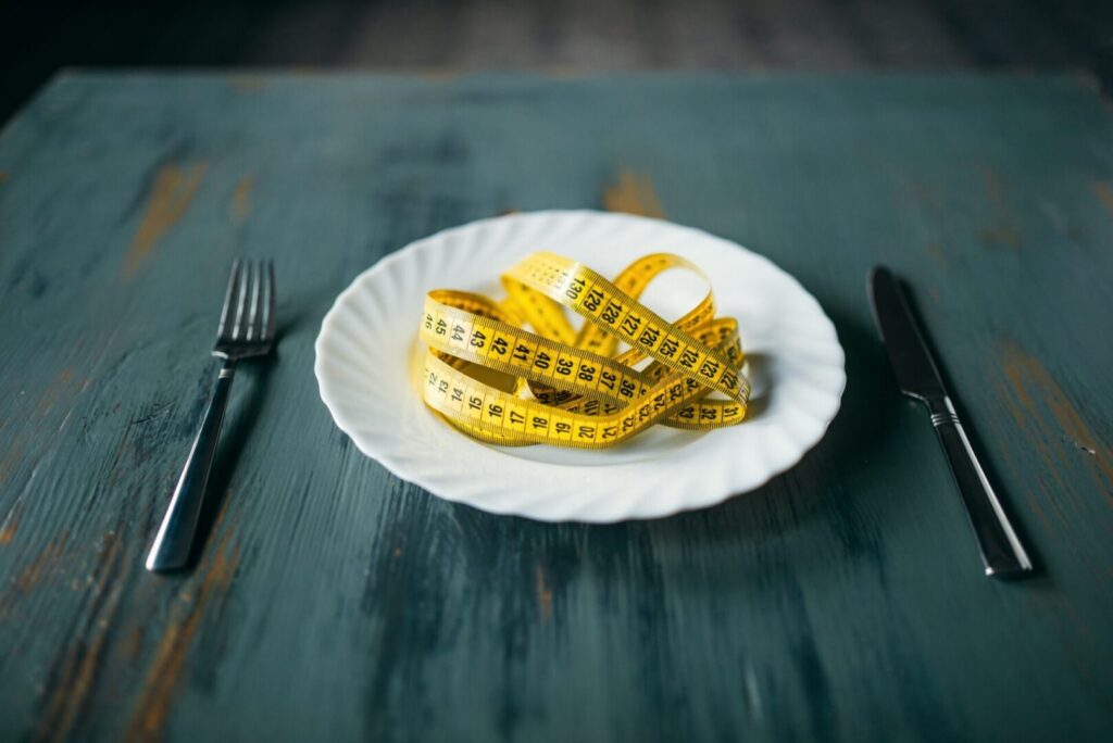 A yellow measuring tape on a plate, with a fork and knife on either side, symbolizes dieting or calorie counting.