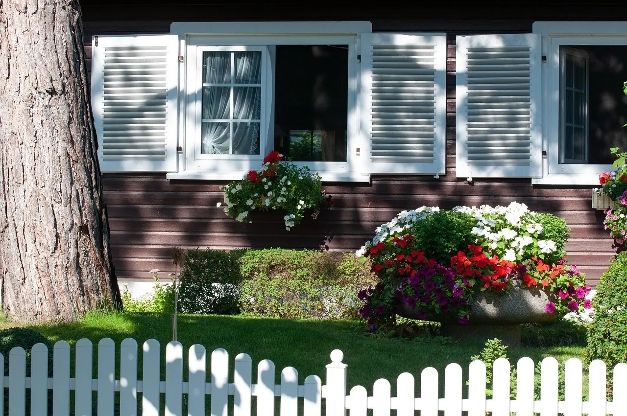 house with white fence A cozy wooden house with white shutters, a tree, vibrant flowers, and a white picket fence.