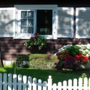A cozy wooden house with white shutters, a tree, vibrant flowers, and a white picket fence.